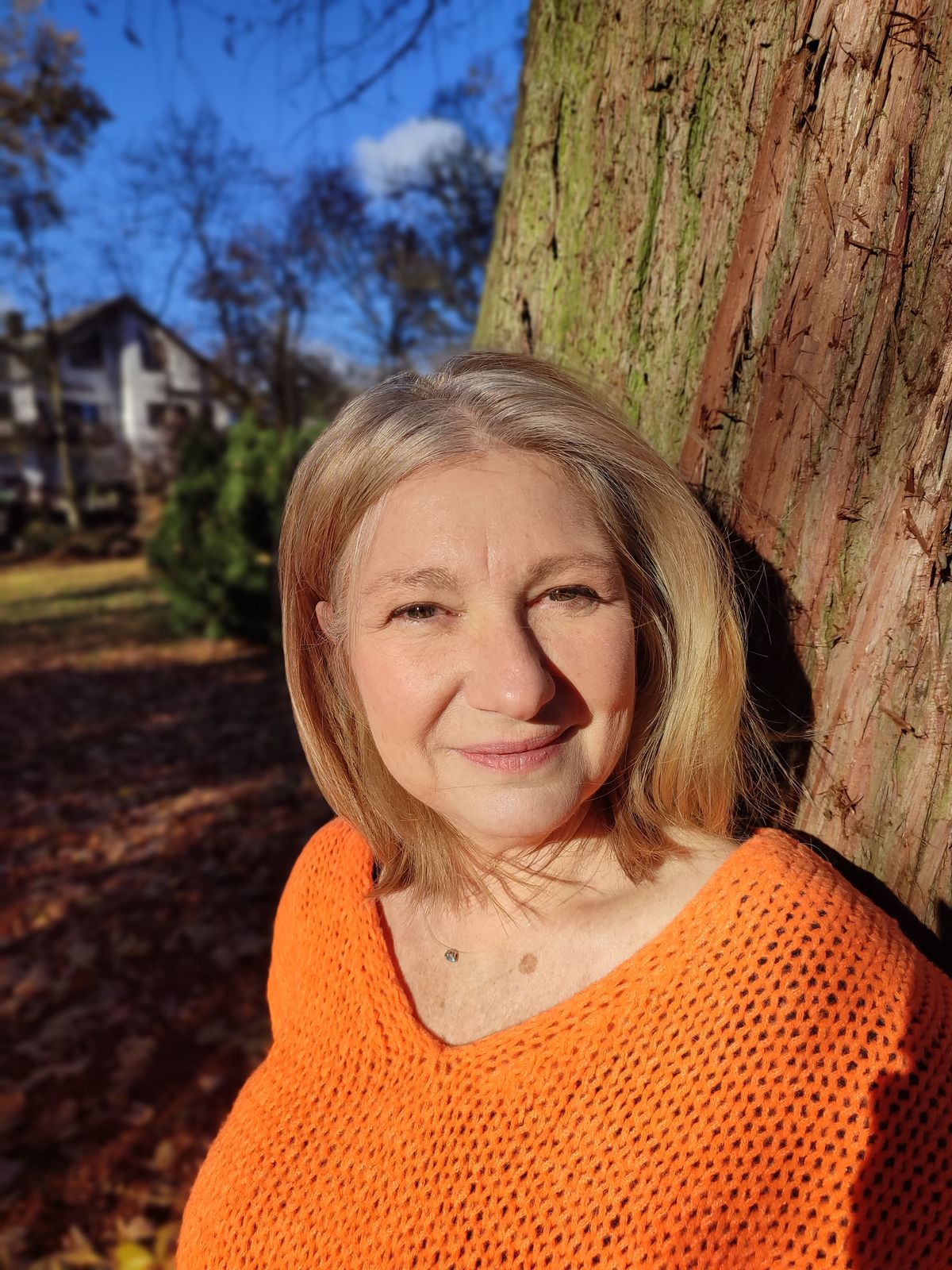 Professionelles Porträt von Beate im orangefarbenen Pullover vor Baumrinde und blauem Himmel